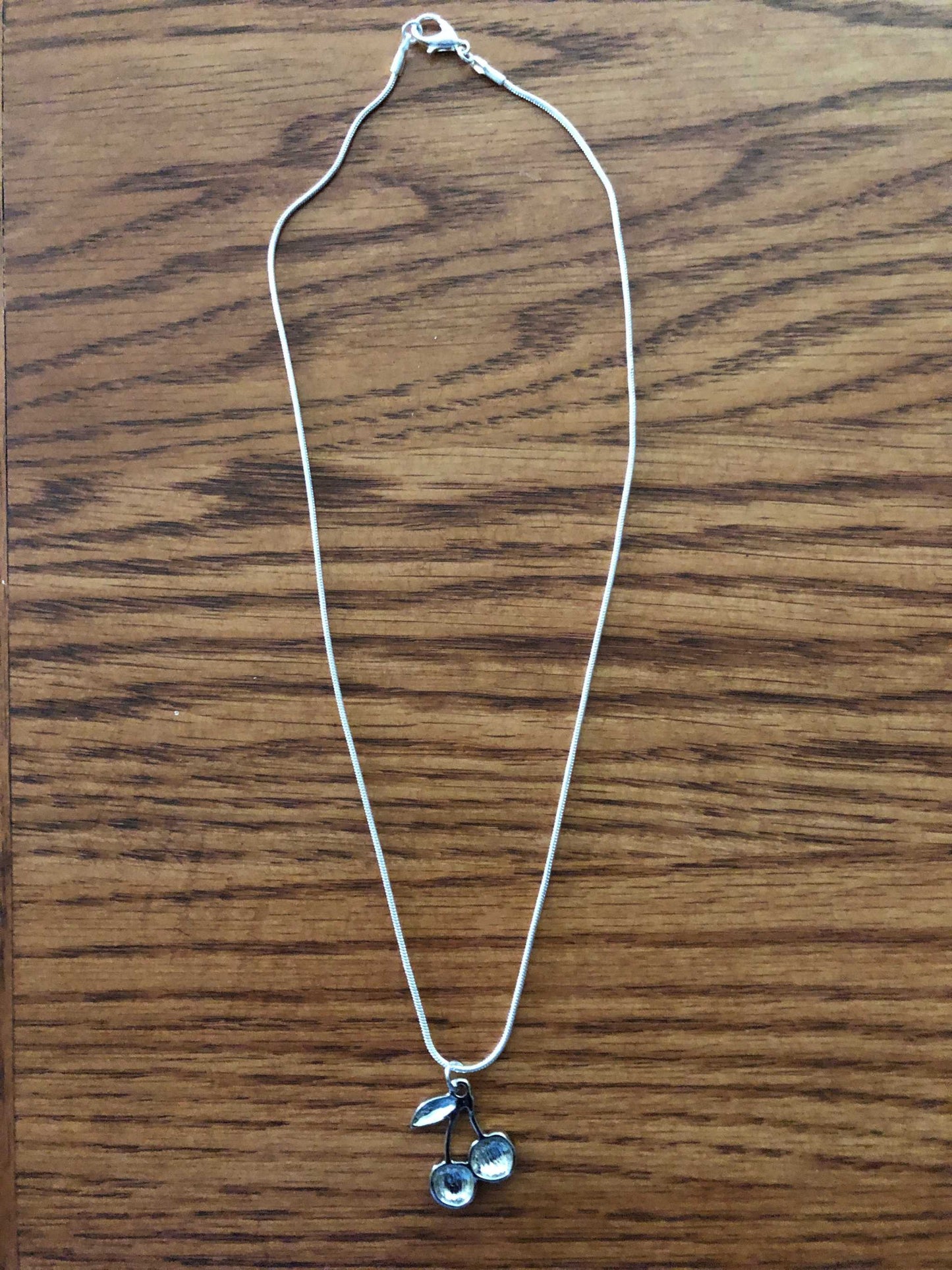 Silver metal cherry pendant on a thin rope chain, displayed on a brown wood surface.