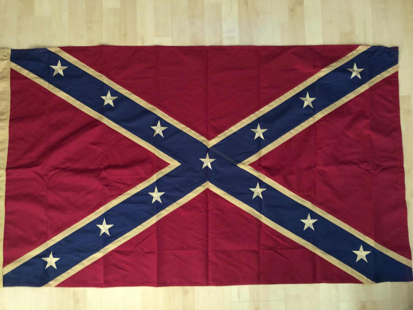 A Confederate battle flag laid flat on a light wood floor, with a red field, blue diagonal cross, and white stars and trim.