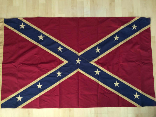 A Confederate battle flag laid flat on a light wood floor, with a red field, blue diagonal cross, and white stars and trim.