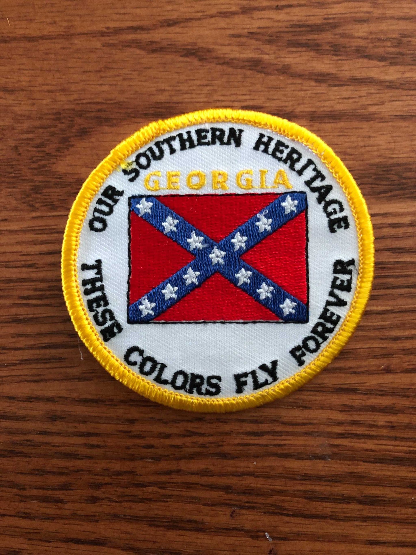 A circular embroidered patch featuring the Confederate flag design with a yellow border and text reading 'OUR SOUTHERN HERITAGE GEORGIA FLY FOREVER THESE COLORS' on a wooden surface.